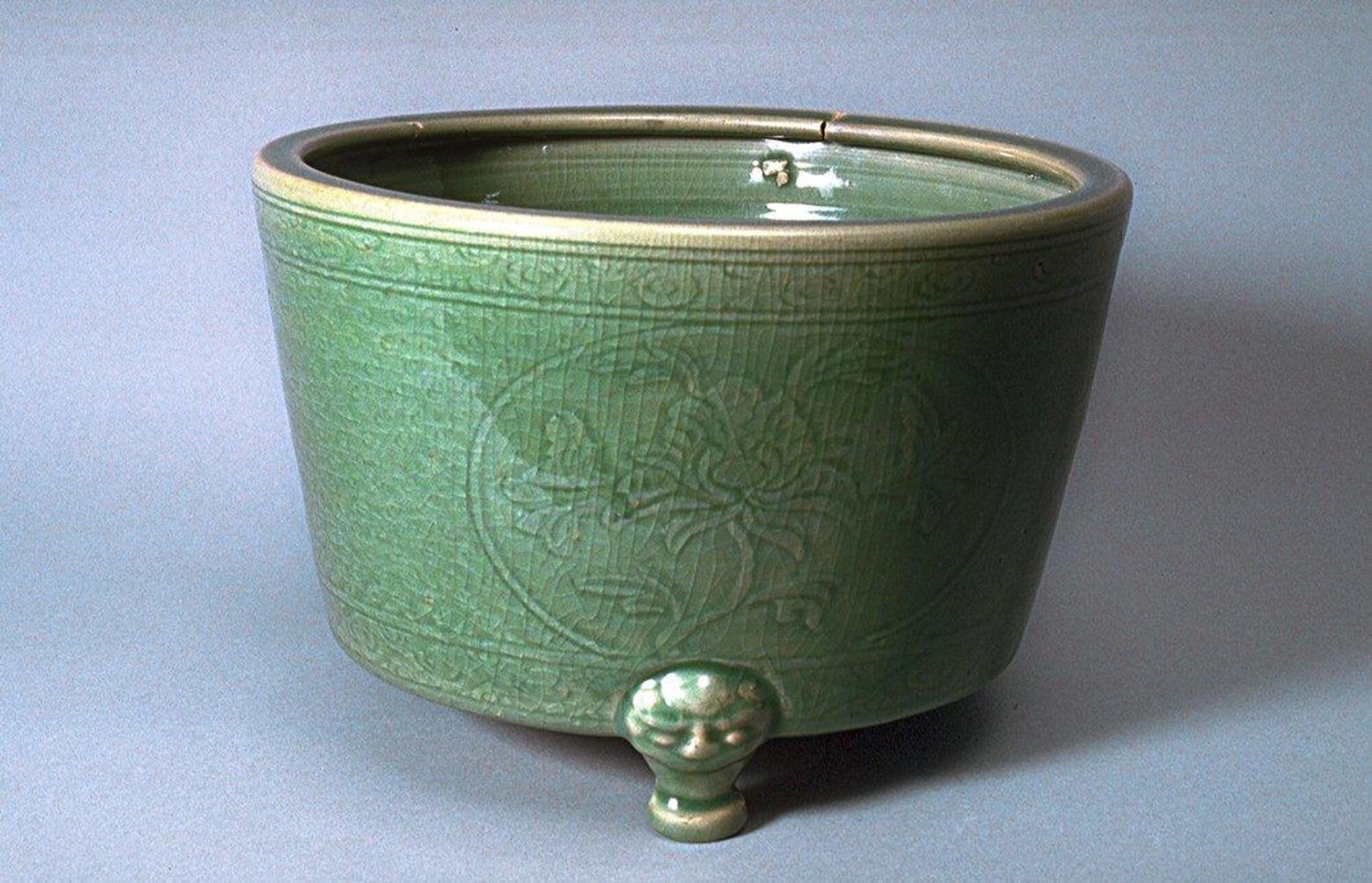 A cylindrical, everted stoneware vessel with animal mask tripod legs applied to the sides.  The body of the vessel is incised with roundels containing peonies surrounded by silk worm scrolls contained between bands of floral meander.  It is covered in a green celadon glaze. 