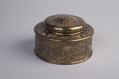 This inkwell has a short cylindrical shape with a circlular hole iin the top and a hinged lid. The body is made of glass with a brass metal overlay decoration of vegetal designs. The lid and ink cup are metal.<br />