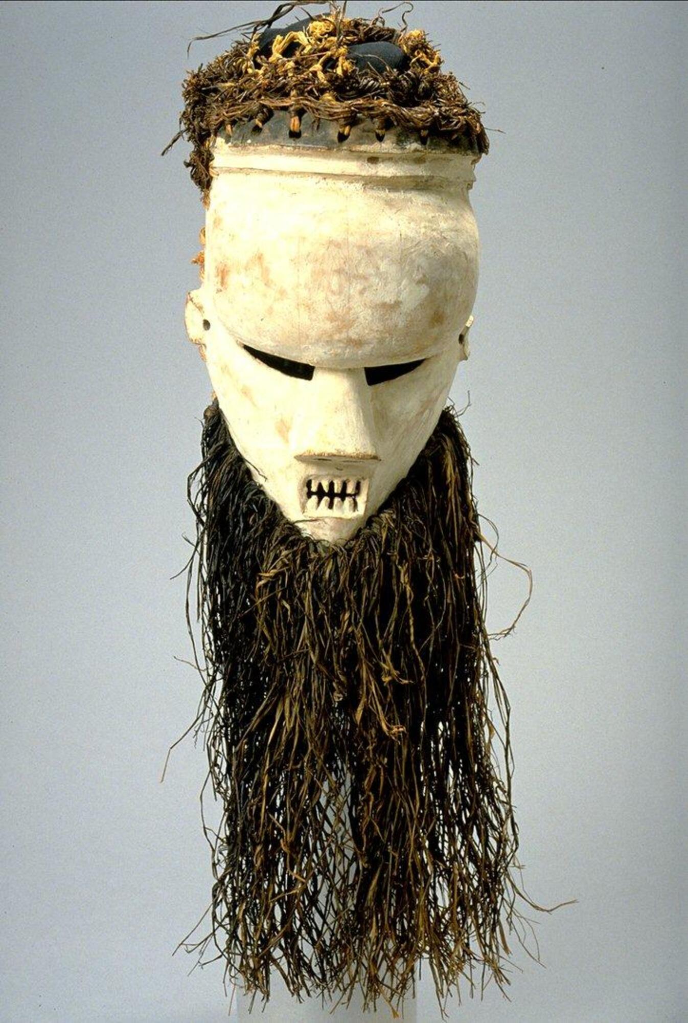 Face mask made of wood, covered in white kaolin; face has round, bulging forehead, deep set narrow eyes, small round ears, fiber beard, open rectangular mouth and pointed teeth; basketry weave that held mask on the dancer’s head is visible at back and sides; raffia attachment on top of head frayed and missing.<br />