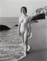A photograph of a nude woman, standing with her feet in the water as it drifts ashore. Behind her, rocks and cliffs are visible in the background.