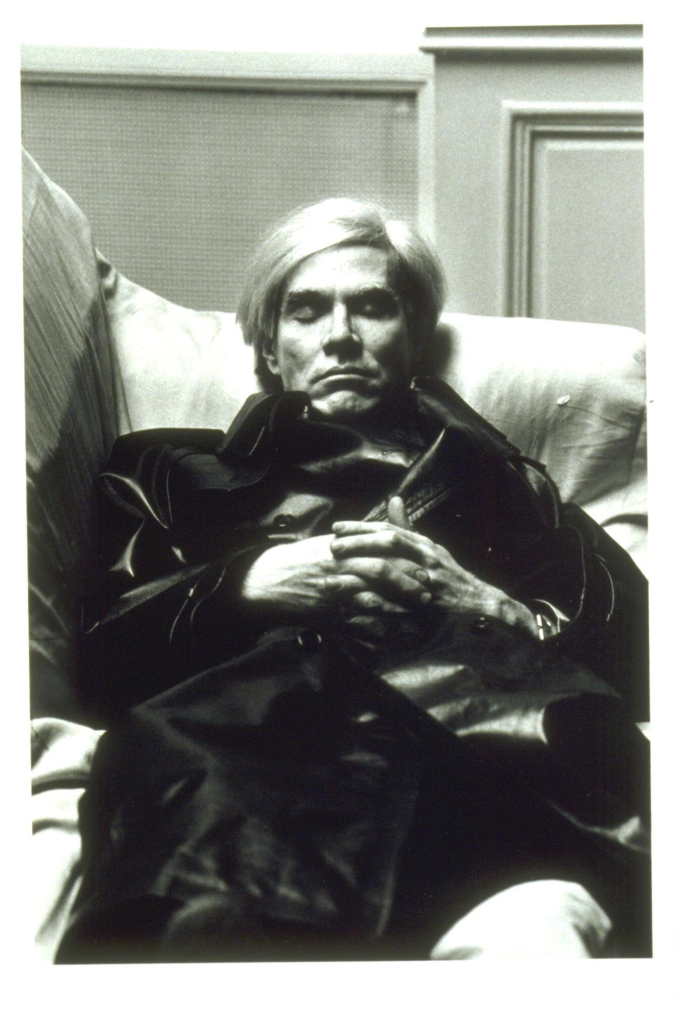 A photograph of a man reclined on a sofa. His eyes are closed and his hands are clasped and resting on his chest.
