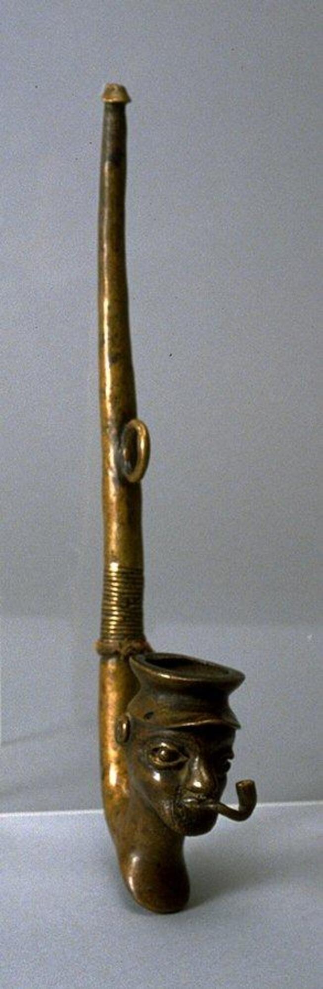 A brass pipe with a figurative bowl in the shape of a human head wearing a cap and smoking a pipe. 