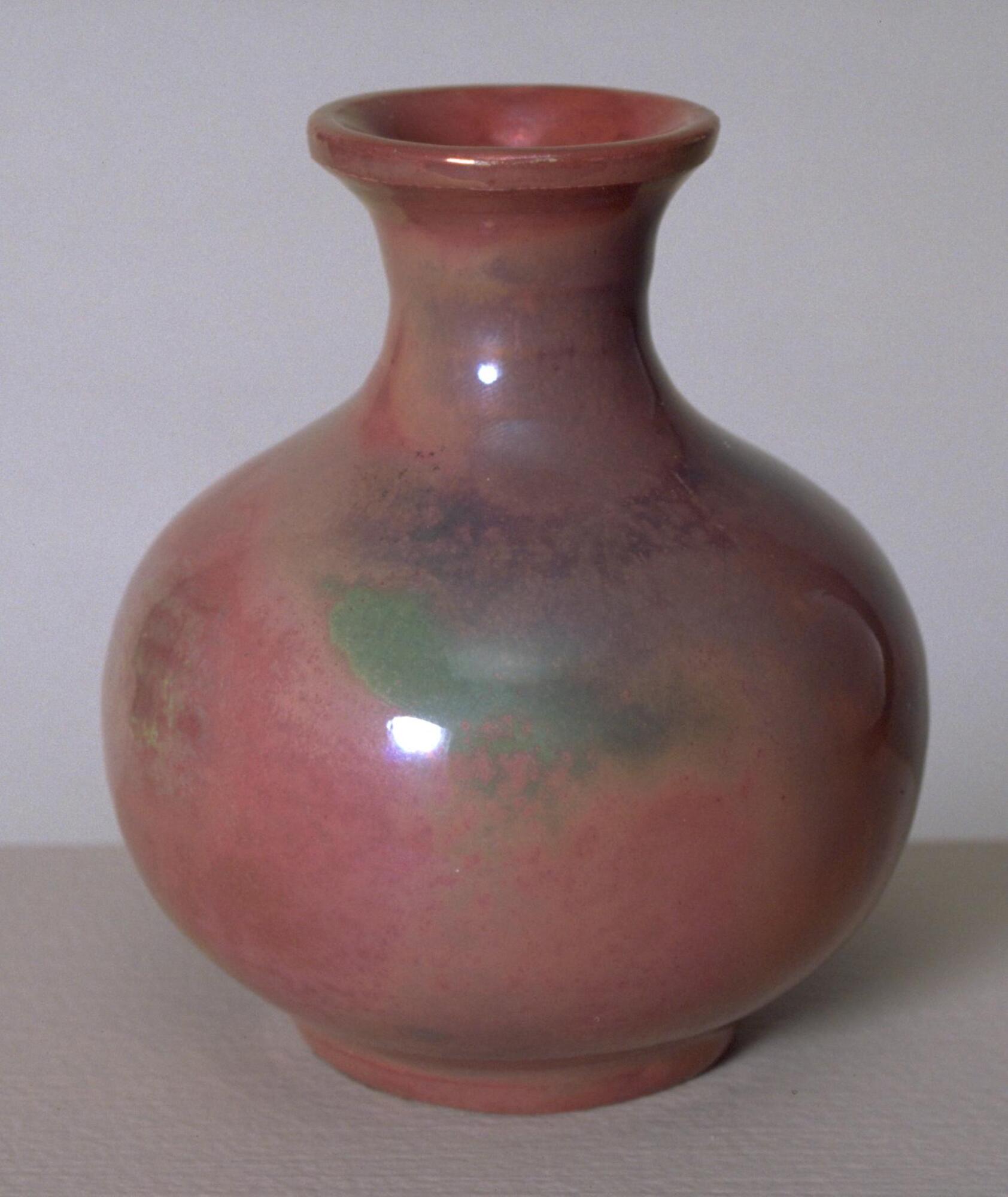 Ceramic bottle-shaped footed vessel with bulbous body, narrow neck and flared mouth covered in an iridescent glaze over a semi-matte glaze that creates an appearance of irregular patches of color in greens and blues with an overall rose color.