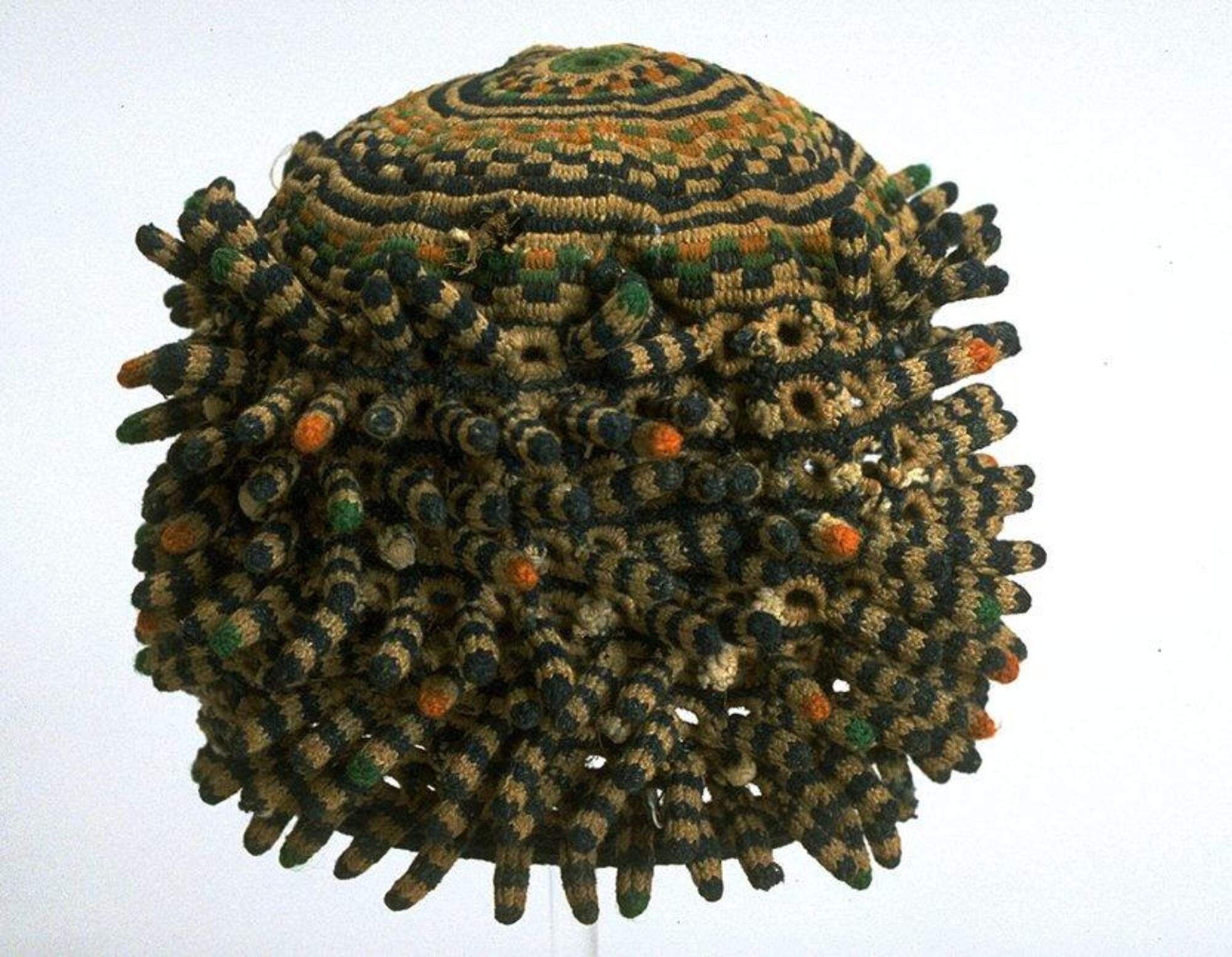This knitted or crocheted hat in a tall, cylindrical shape has concentric circles and checkerboard patterns decorating the top. The body of the hat is made of small open circles and short rod-like projections. 