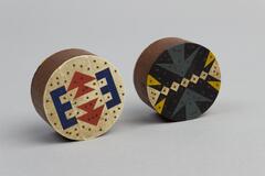 Each short wooden cylinder is painted with an inlay on both circular face. One face is red, white and blue with brown dots. Other face in dark teal, black, yellow and white with brown dots.