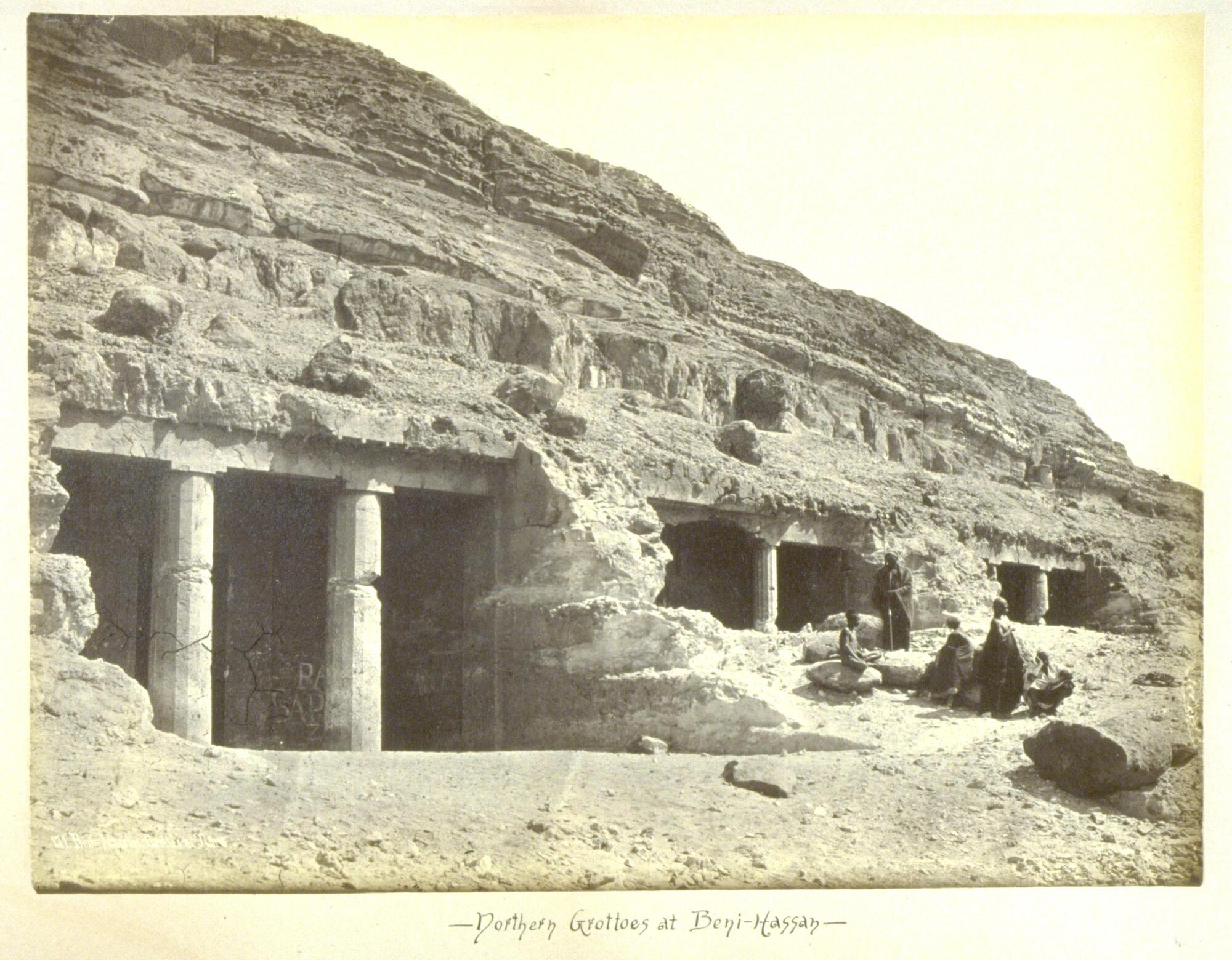 Five men sit just outside the rectangular, columned entrances of three grottoes set into the side of a hill. 