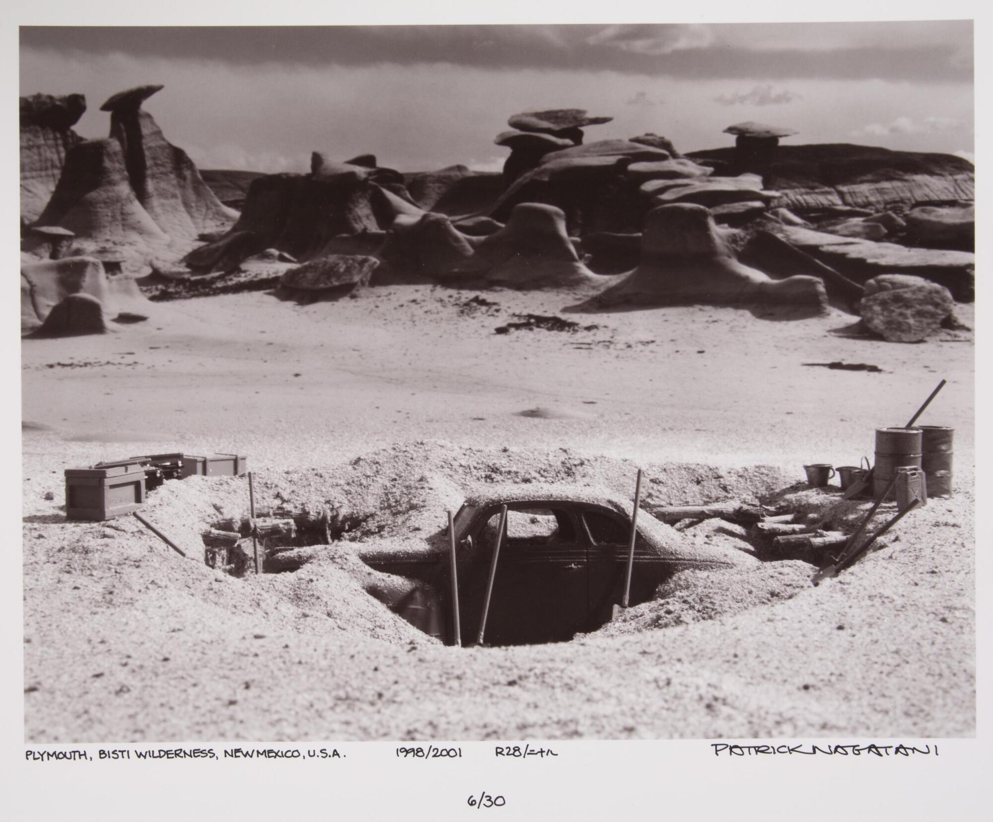 This photograph depicts a view of a sandy desert with eroded rock formations in the background. In the foreground is an archaeological excavation of a buried car.