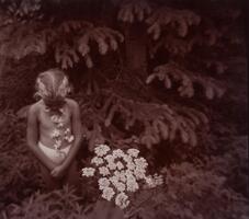 In this photography, a young girl stands in front of a large coniferous tree with plants surrounding her. In the foreground is a large cluster of white flowers. She looks down, wearing a string of flowers around her neck with her hands clasped in front of her.