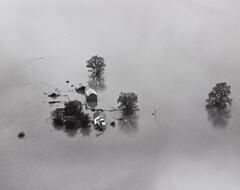 An aerial view of a flooded farmhouse, outbuildings and trees with boats at the farmhouse door.