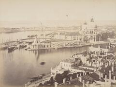 This photograph depicts a wide, ariel view of a city situated on thin strips of land with wide canals running between them. On the central island is a large white church and a wharf with a docked fleet of boats.