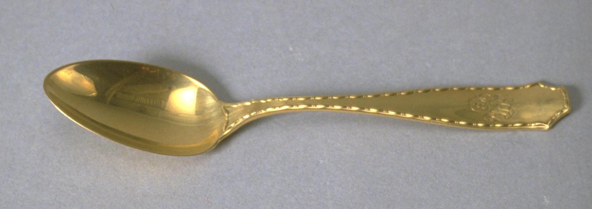 Gold spoon with thin handle that widens at the end with egg-and-dart-like motif along edges