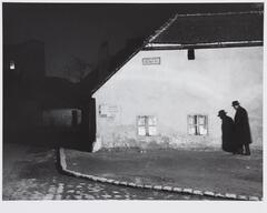 Photograph of a man in an overcoat and hat walking close to the side of a house, with his shadow projected on the wall beside him.
