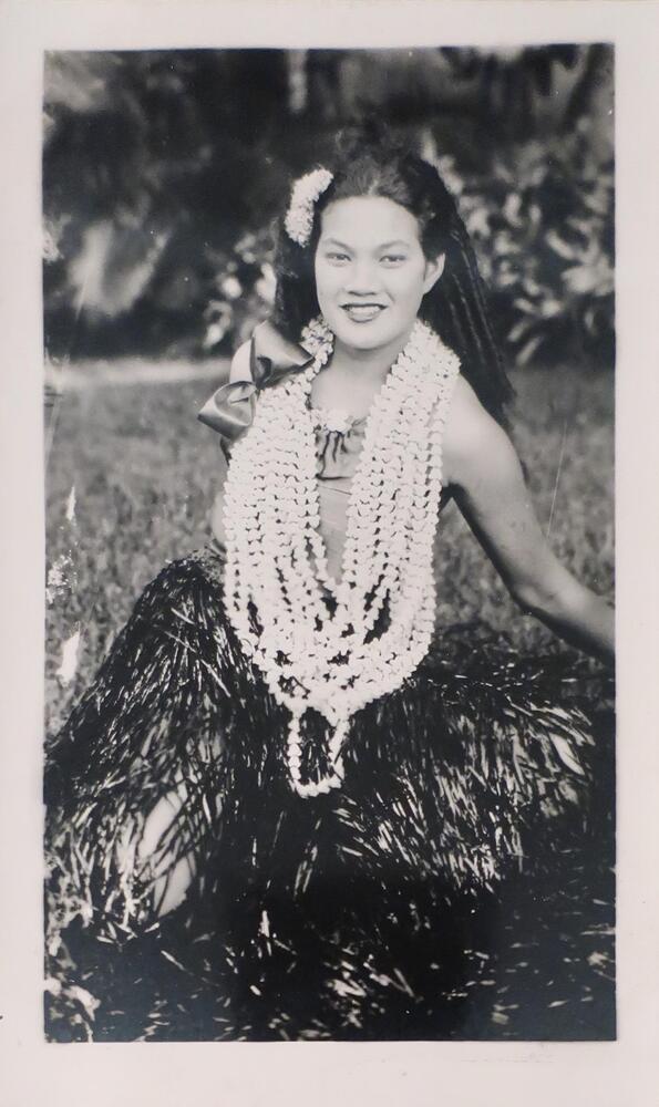 A smiling woman in a grass skirt and multi-string necklace sitting on the grass. She has flowers in her hair and a large bow near her shoulder.