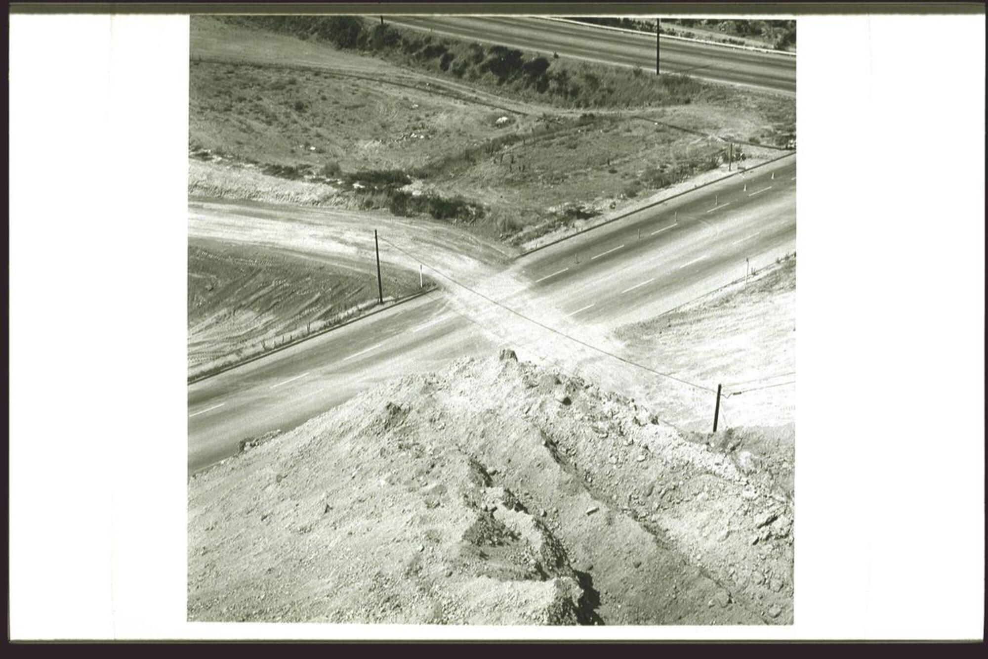 This is a black and white photograph of some roads seen from above, as if the viewer is standing on a nearby hillside. In the foreground, at the viewer's feet, is rocky, dusty soil. Below, the main road runs diagonally across the work and a smaller dirt road crosses it, leaving traces of dirt tracks to the other side of the road. The land is barren with very little vegetation and no vehicles are to be seen.<br />