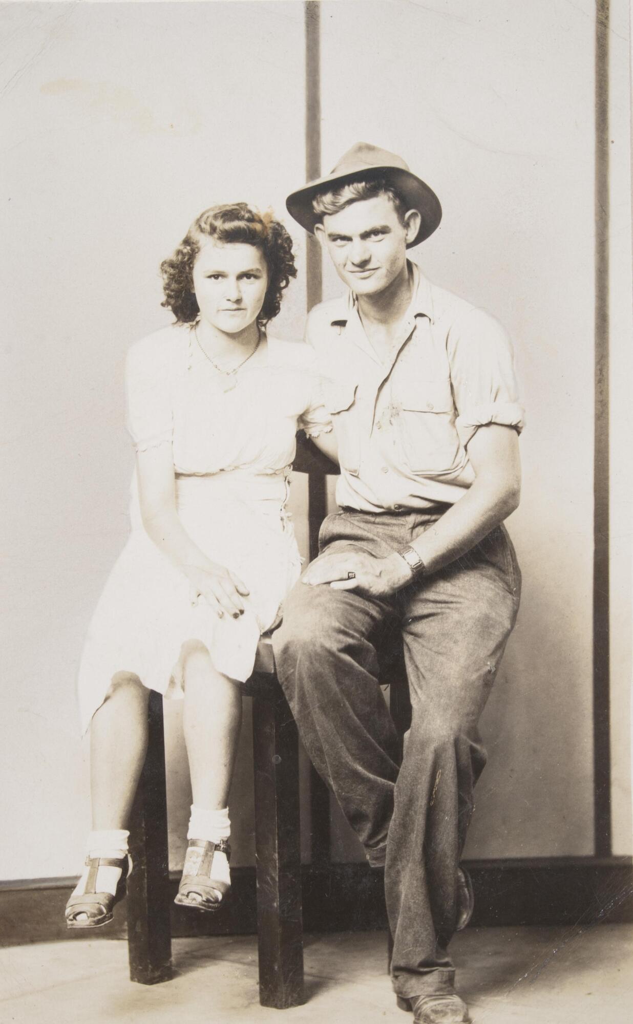 A couple seated in front of a striped backdrop with their arms around one another. A woman sits on the left, wearing a light dress, her feet hovering above the ground. A man sits on the right, wearing trousers, a light short-sleeved shirt, and hat.