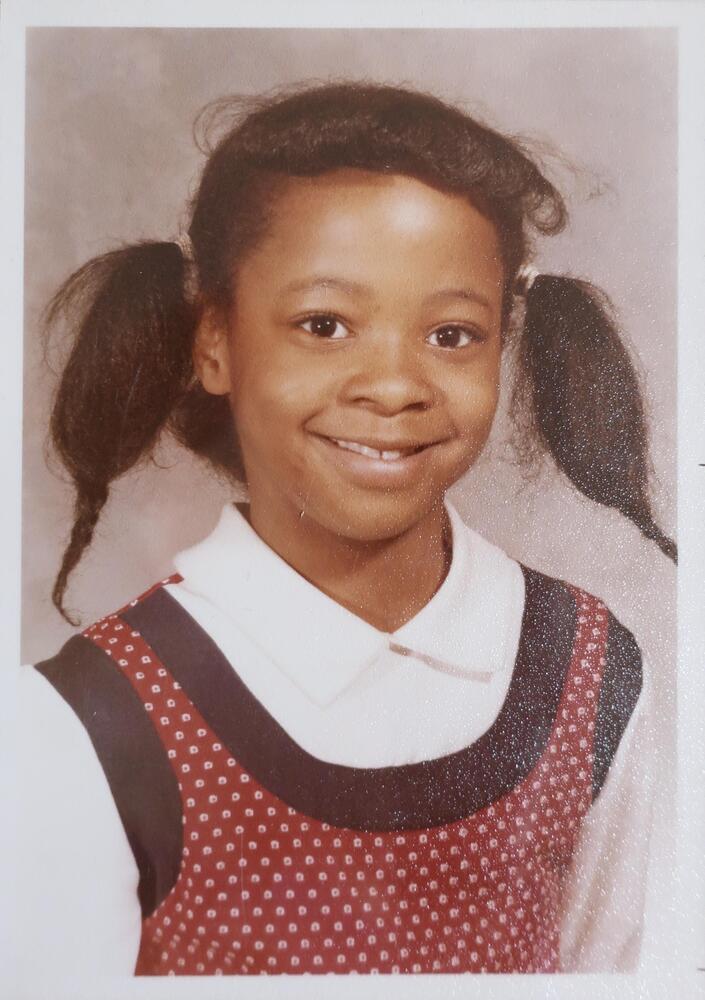 A portrait of a smiling young girl with pigtails. She wears a white collared shirt underneath a spotted red jumper and her pigtails end in short braids.