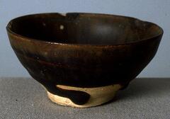 A bowl on a footring, covered in a thickly applied, iron-rich black glaze. The bottom of the teabowl is not covered with the glaze except where the glaze pooled in certain places.