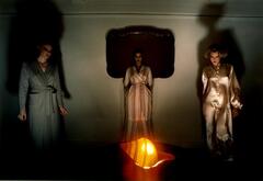 This work is a color photograph of three models in night wear. One woman stands in the center background, while two models stand on the far sides of the frame, their shadows elongating toward the ceiling. Each looks at, and seems to move toward the center, where an illuminated conch shell glows on a table.