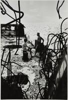 A sitting woman and standing man face each other on the surface of a wrecked structure at the side of a large body of water. Curling metal bars burst from crumbling concrete walls in the foreground.