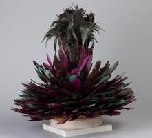 A headdress made of woven raffia palm fibers in the shape of a cap and decorated with dark purple and green feathers. The feathers point upward and outward from the cap, while the center of the headdress has a bundle of large, dark feathers. 