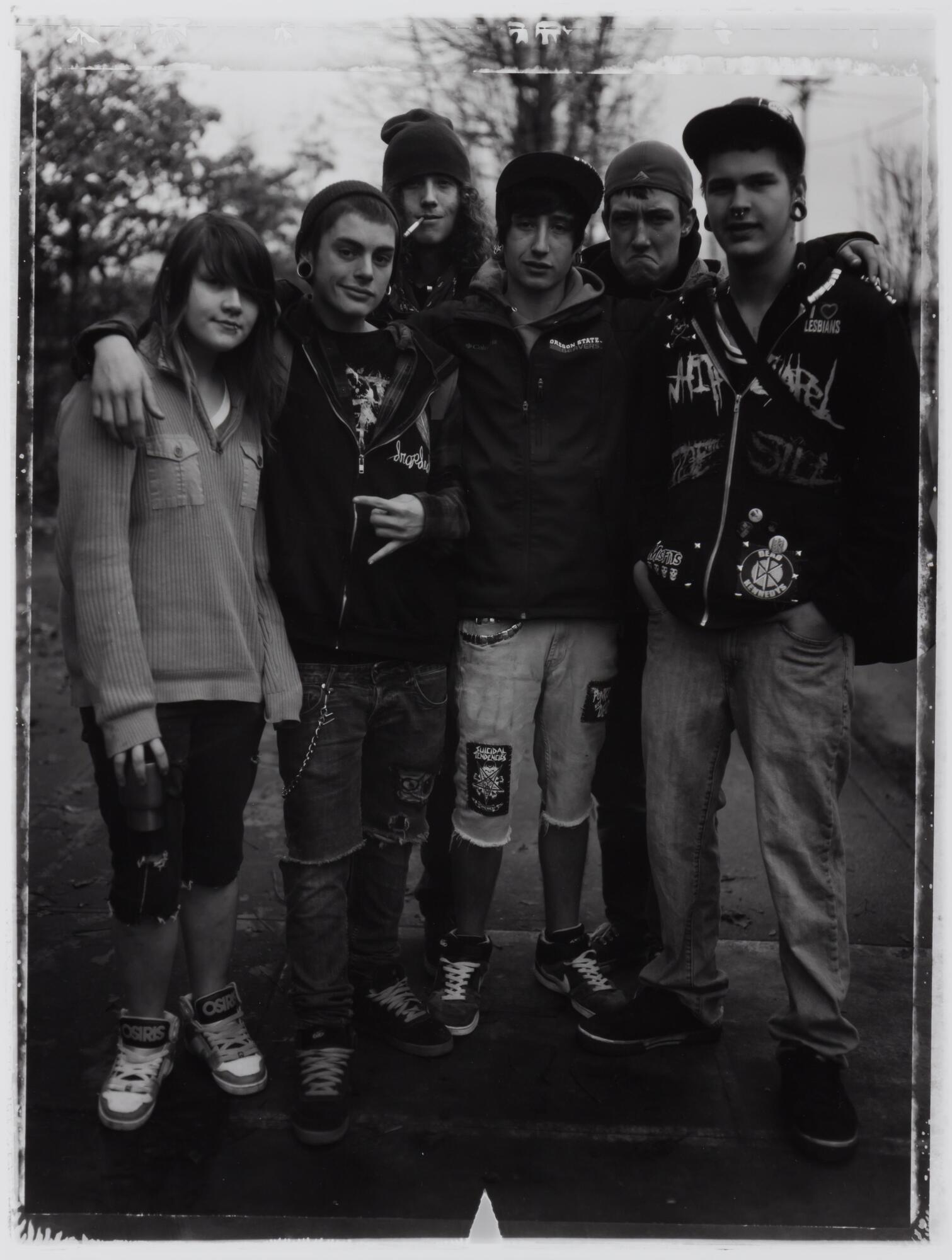 This photograph depicts a group of teenagers wearing punk rock-style clothing with their arms around one another and posing for the camera. 