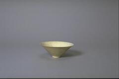 This thin porcelain conical bowl has a direct slightly everted rim on a footring. Its interior has a lightly incised floral meander decoration,and it is covered in a white glaze with bluish tinge.
