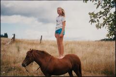 A blonde girl standing on top of a horse in a field.