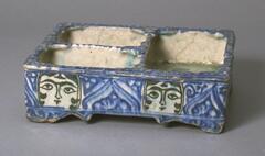 This Qajar dish features three separate compartments and highly decorated exterior panels. Each side of the dish is decorated with a pair of young female faces that alternate with abstract deep blue designs. The interor panels lack decoration aside from the bases of each compartment which contain blue painted floral sprays. The craftmanship of the dish finds roots in the Kashan tradition of the 12th and 13th centuries, making it a testament to the continuation of traditional techniques in the region by the 19th century. 