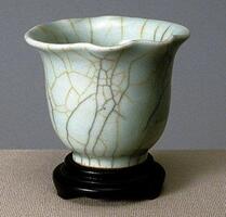 A porcelain, bowl-shaped cup on a tall foot ring with a flaring, undulating rim. It is covered in a crackled celadon glaze.&nbsp;