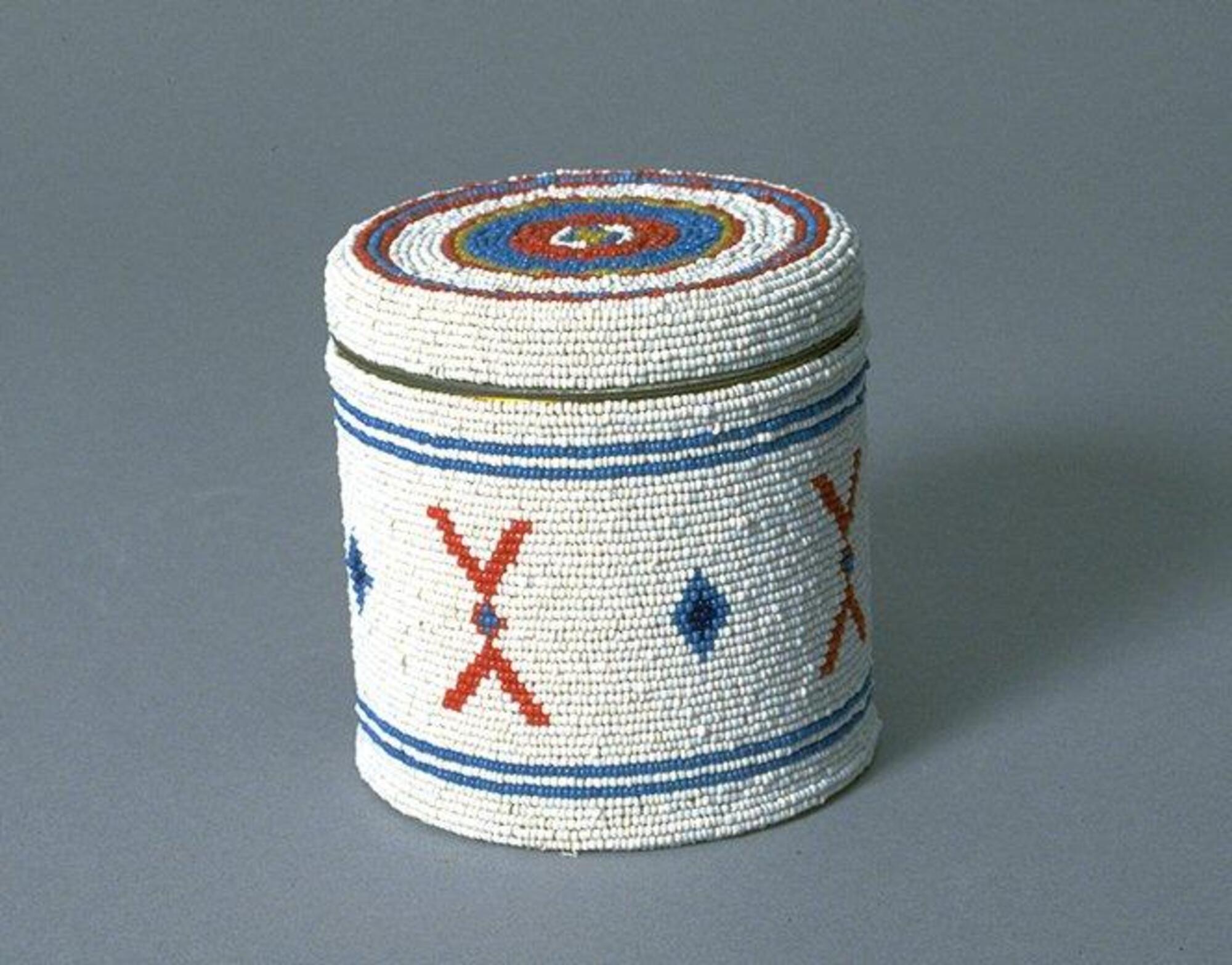 Cyllindrical metal container decorated with glass seed beeds: white ground with blue stripes and diamonds, red X's, and concentric circles. 