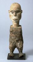 Standing figure on a square base. The head is large with a high, rounded forehead and wide cheeks. The almond-shaped eyes sit below slightly curved eyebrows. The mouth is closed. The neck sits atop a columnar body without arms. The legs are short and in an open stance. 