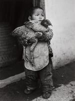 A photograph of a small, young boy in soiled clothing. The boy holds a chicken in his arms and stands in the street, just outside of a building.