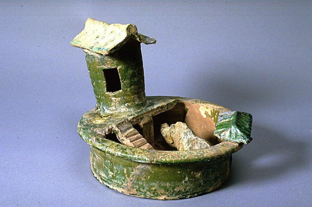 A red, circular-shaped earthenware pigpen, containing one pig, below a cylindrical tower shed with a window and a peaked roof displaying ridges.  Stairs connect the shed to the pen.  It is covered in a green lead glaze with iridescence and calcification. 