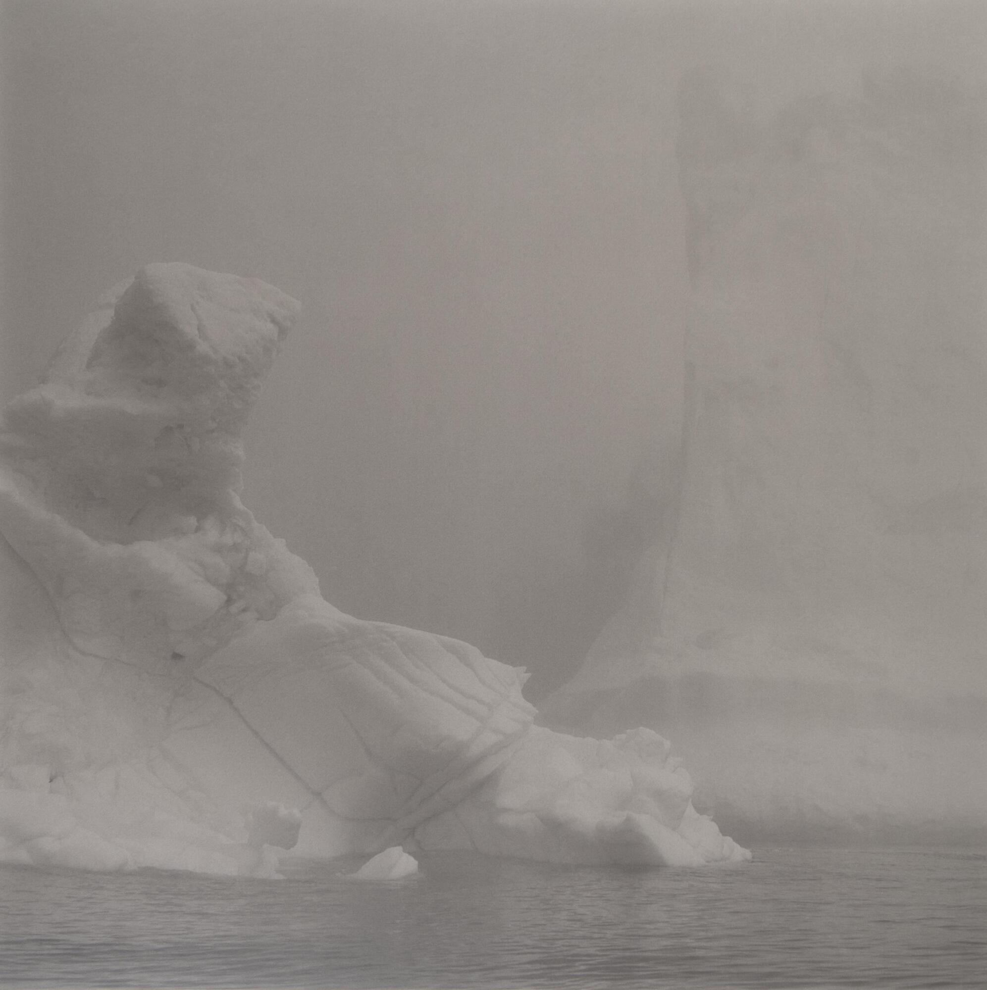 This photograph depicts an iceberg in a bay. The iceberg fills most of the frame and subtely emerges from the background.