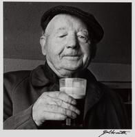 An older man with a knitted cap, holding up a pint of beer. He is wearing a dark top and the background is dark.