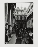 People fill a narrow street lined with tall buildings. Three women walk towards the camera smiling to each other.