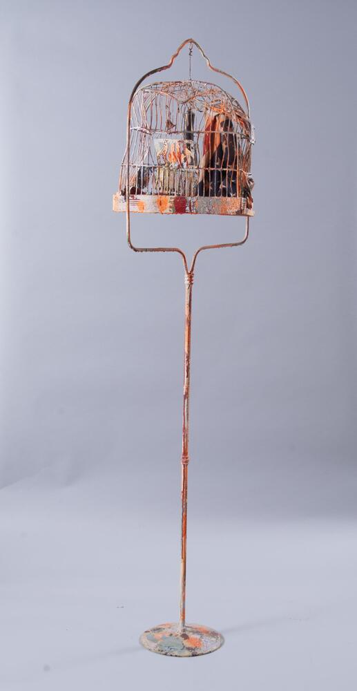 A splatter painted bird cage stand with a rounded top cage on top in red, green, orange, white and blue. There is mesh on one side of the bottom of the cage that is also splatter painted. Inside the cage hanging from the top is a small bell, part of an American flag and a replica of a human penis, painted black, hanging upside down.
