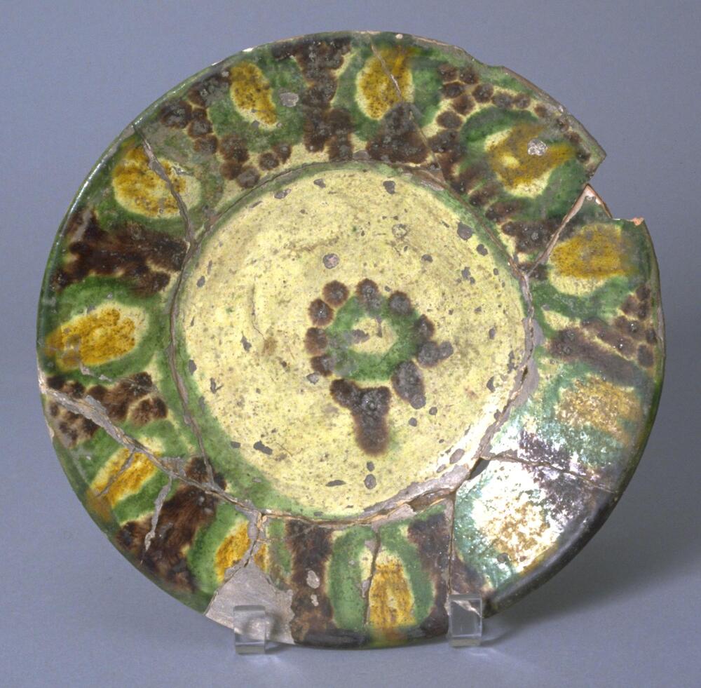 'Ueberlauf' on rim running design, in center a blossom; paste: buff, fine, medium-hard; glaze: glossy, fine crackle top glaze, over cream slip on interior and exterior except bottom part. Fired upside down (tripod on interior) and upright. Colors are green, yellow, aubergine, green-white. Slightly restored.