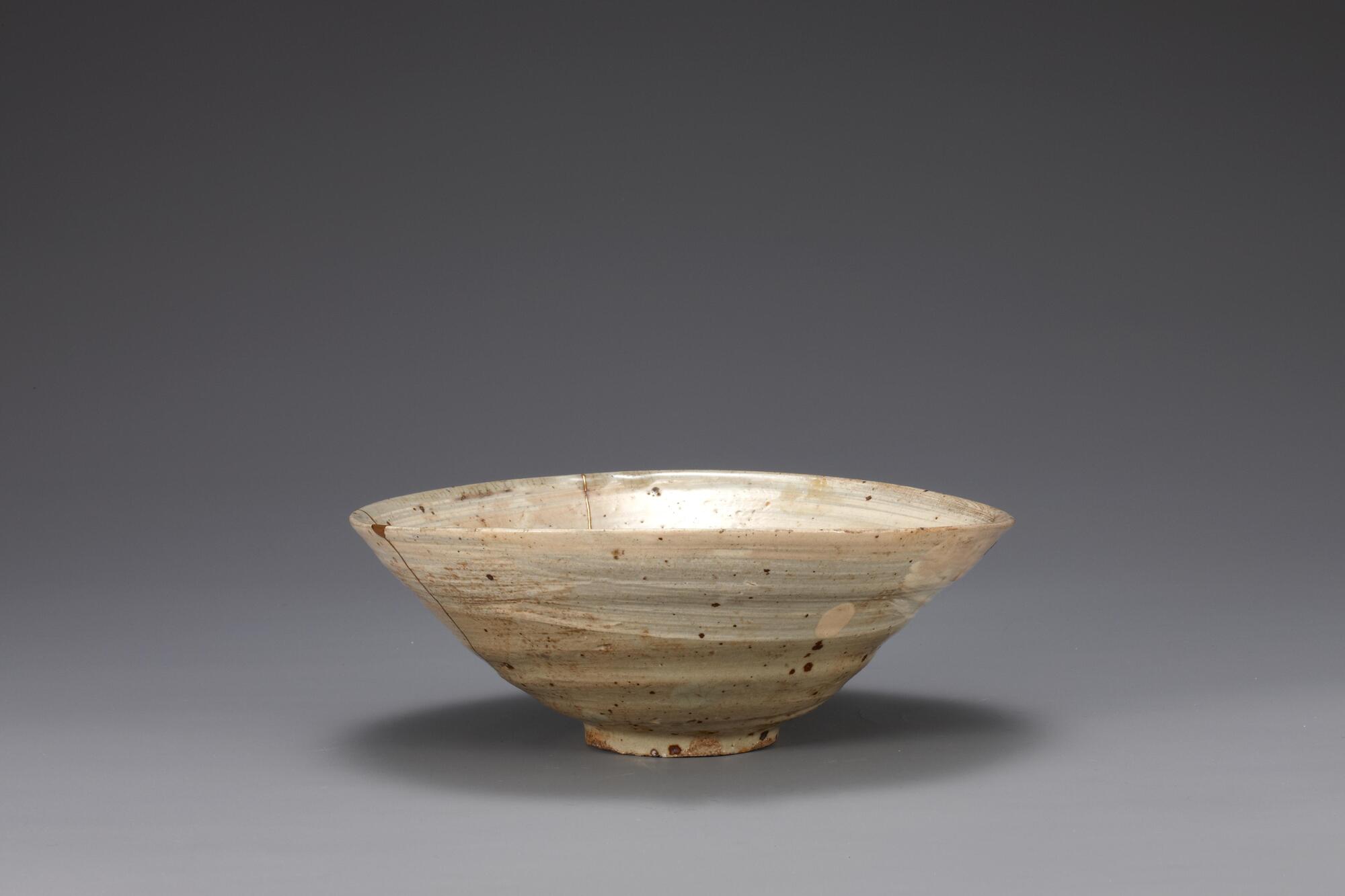 It has large-mouth and the wall slants toward the bottom in an almost straight line. It was deformed during firing.<br />
<br />
The inner and outer surfaces of this bowl are brushed with white slip. Traces of refractory spurs remain in five places on the inner base and the foot. The foot is glazed on the rim as well. This is a low-grade object with many iron-brown spots on both its inner and outer surfaces. A note from collector is adhered to the reverse side of the bowl, and it reads, &ldquo;19629, cor&eacute;e, Hak&eacute;me,&rdquo; &ldquo;朝鮮刷毛目, 三百年.&rdquo;<br />
[Korean Collection, University of Michigan Museum of Art (2014) p.156]