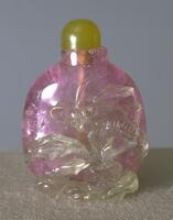 A pink tourmaline snuff bottle with white tourmaline raised relief carving of a grasshopper and grass. On top of the snuff bottle is a stopper of serpentine.