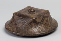 A lid for a square shaped box with a rounded disk-shaped base. The lid is bordered with a diamond geometric pattern and the center has a round button-like fixture attached to it. For a full description of the box, please see object no. 1985/1.162B.