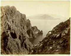 This photograph depicts a view from a rocky coastline looking back at the mainland coast. In the right portion of the frame, a woman in a dress sits on a boulder and looks out to sea.