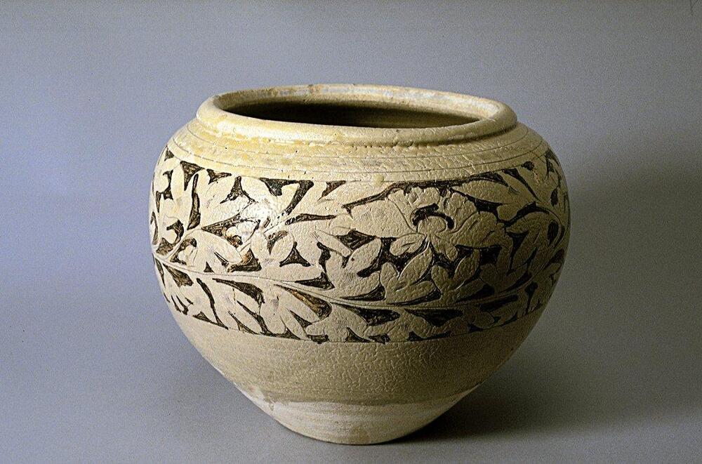 A stoneware globular jar with wide mouth and belly tapering towards the foot. The jar is covered in a white slip, carved with floral meander, and painted in the recesses with a dark under glaze. It is covered in a translucent glaze.