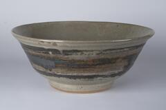 Large, deep and wide ceramic bell-lipped bowl. It sits on a narrow base and curves outward. It is a greenish-gray color; the outside is streaked ith copper and dark brown colored streaks. Celadon glaze with crackle on the inside, with temoku register on the outside; red-color clay.