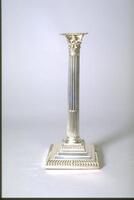 This silver candlestick, one of a pair, is in the shape of a fluted column topped by a composite capital composed of a double row of acanthus leaves and four volutes at the corners. The column rests upon a stepped base made of four squares of diminishing size, the first and fourth of which are marked with a pattern of strigillations.