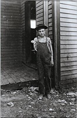 This photograph shows a young boy in overalls and standing before a wooden house missing his right arm.