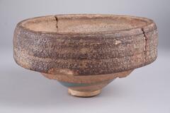 This conical bowl on a straight footring is covered in a pale blue opaque glaze with a copper purple splash to the interior rim and side-wall. It is fused to the stoneware saggar in which it was fired, with cracks to the saggar.