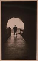 A silhouette of a man, photographed against what appears to be the opening of a tunnel with railings on both sides. Most of the image is very dark with a light circular shape and the figure at the center.