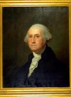 Portrait of George Washington, grey hair, solemn face, wearing a suit with a white shirt underneath.