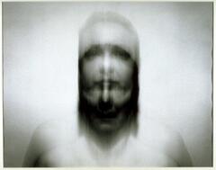 This photograph is a blurred and distorted portrait of a figure standing against a blank wall.
